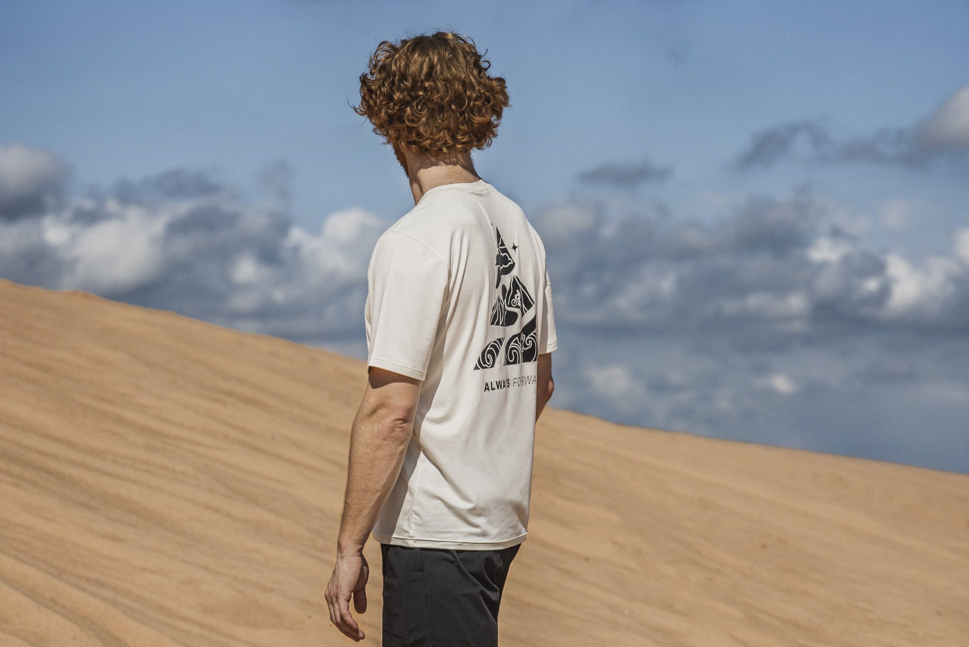 Person standing on a sand dune with a blue sky and clouds in the background