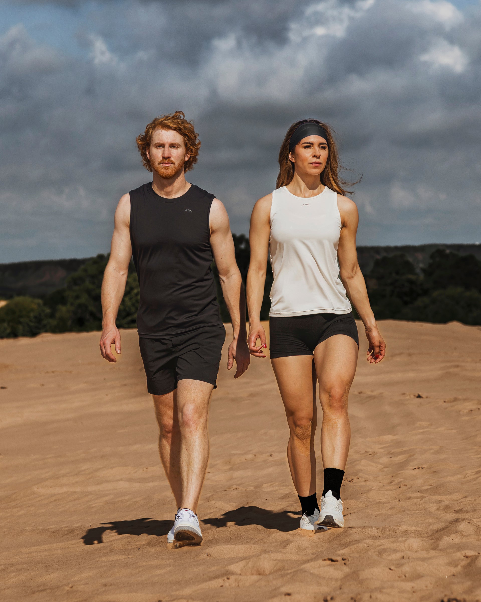 Two people sporting JUNK apparel walking on a sandy landscape with a cloudy sky.