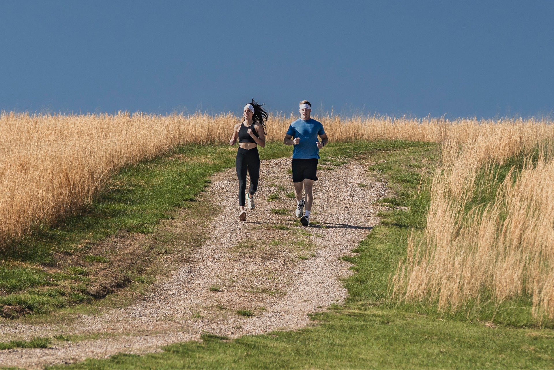 Two people running on a path through tall grass under a clear blue sky.