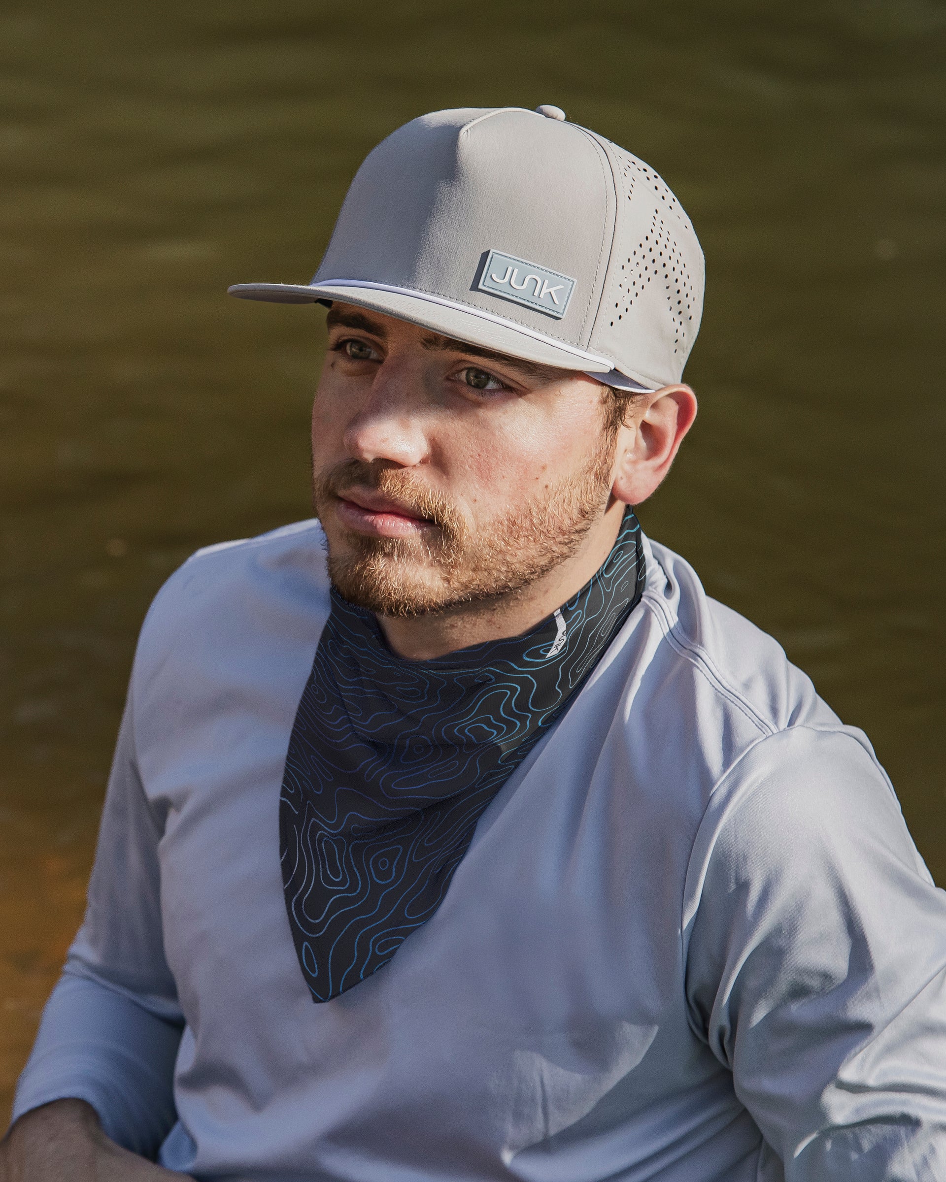 Man wearing a gray Apex Fog hat, a Trail Blazer summer neck gaiter and a light gray shirt with a blurred natural background.