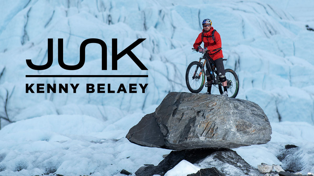 Kenny is on top of a rock with his mountain bike in a red coat in an iced geographic area with text overlay junk logo and “kenny belaey” 