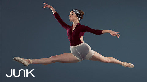 Woman in maroon leotard and white skirt and point shoes and junk headband performing a dancer leap 