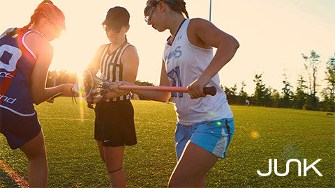    Image in a lacrosse field at sunset, two competitive athletes and a ref about to start the game. The junk logo in bottom right corner 