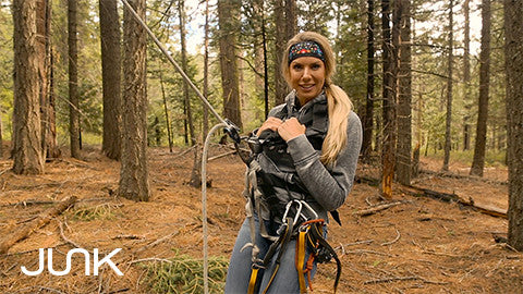 Woman in harness with long blonde ponytail prepped to swing in the woods