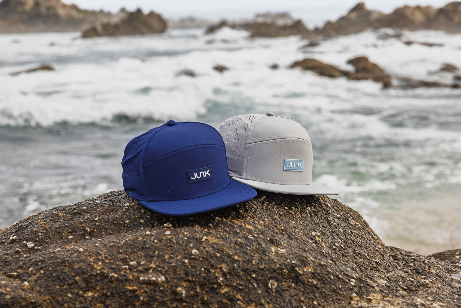 Two caps, one blue and one gray, on a rock with a coastal background.
