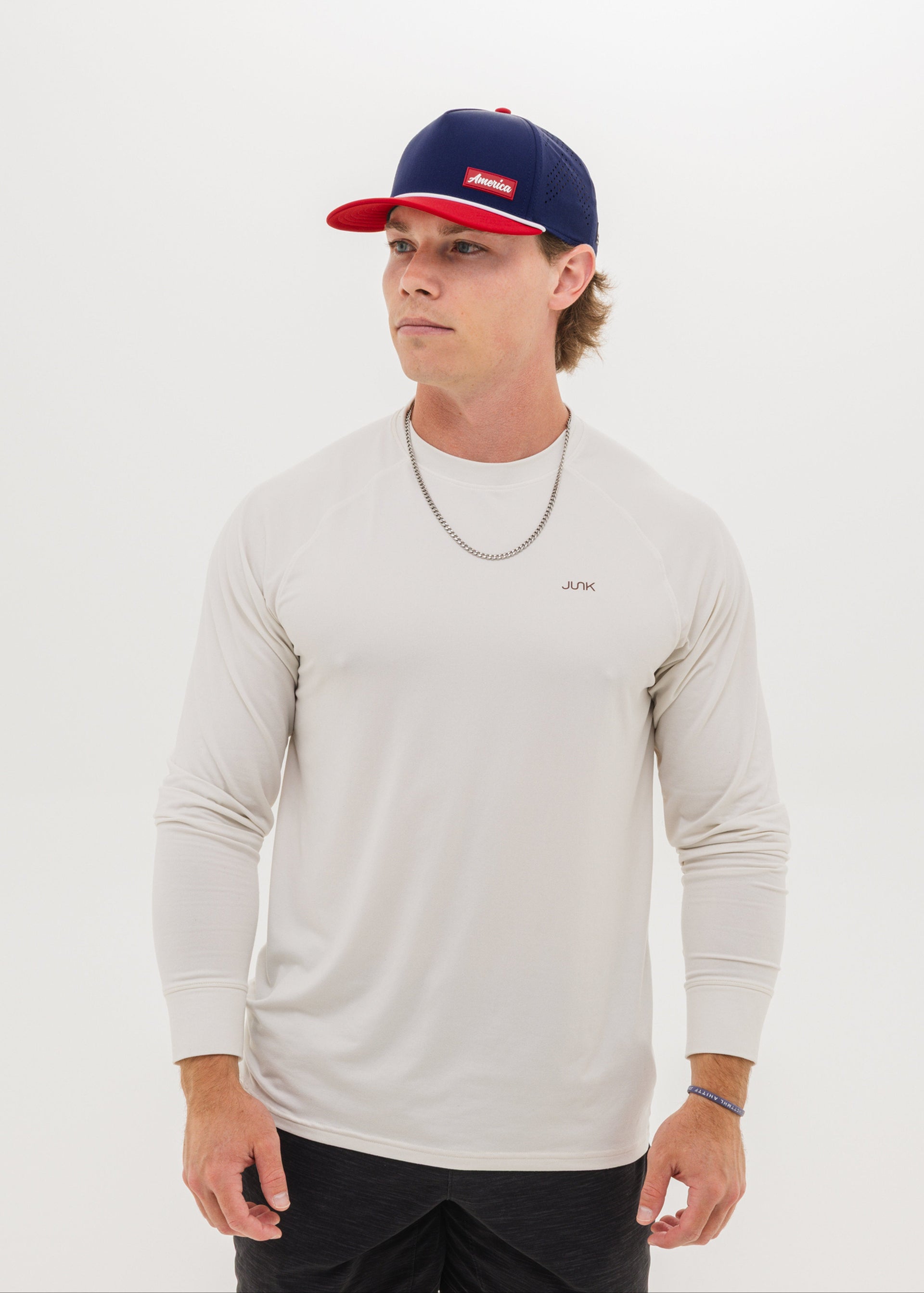 A man wearing a light beige long-sleeve athletic shirt, a navy and red baseball cap, and a silver chain necklace, standing against a plain white studio background.