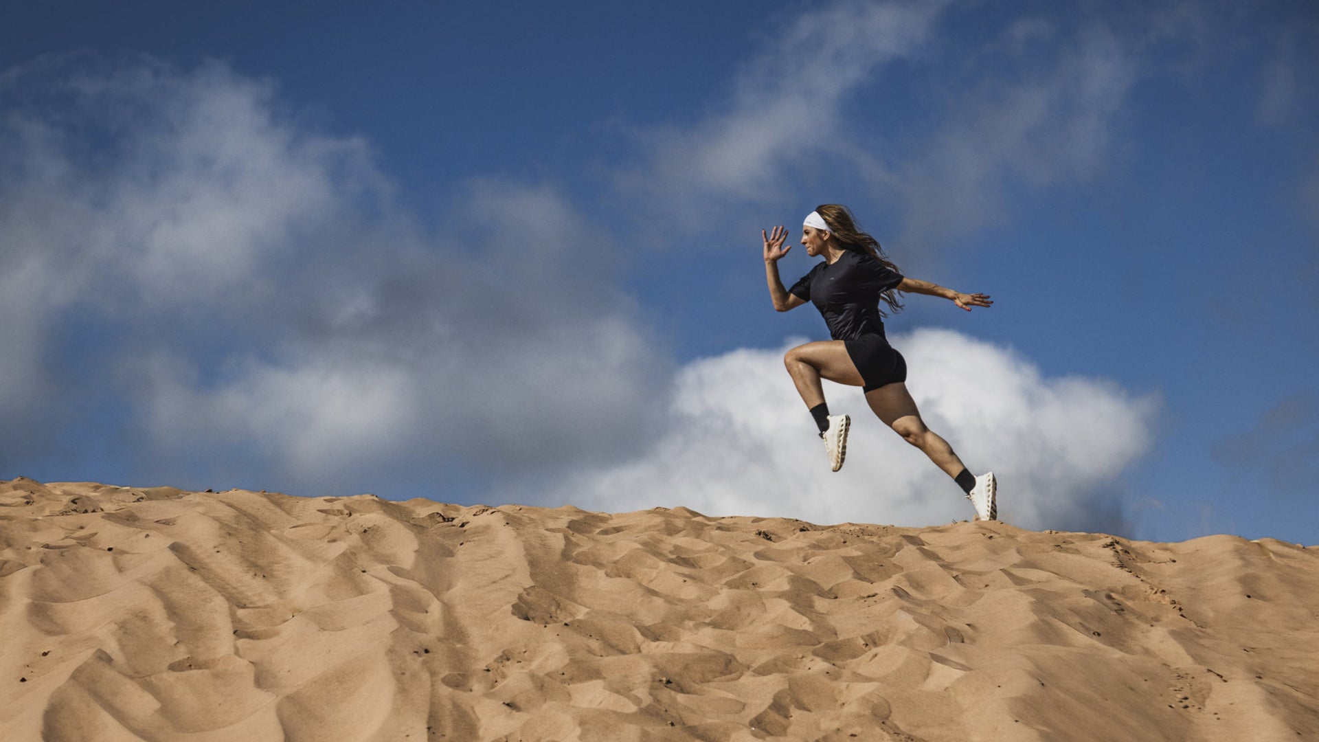 Person running on a sand dune with a blue sky and clouds in the background