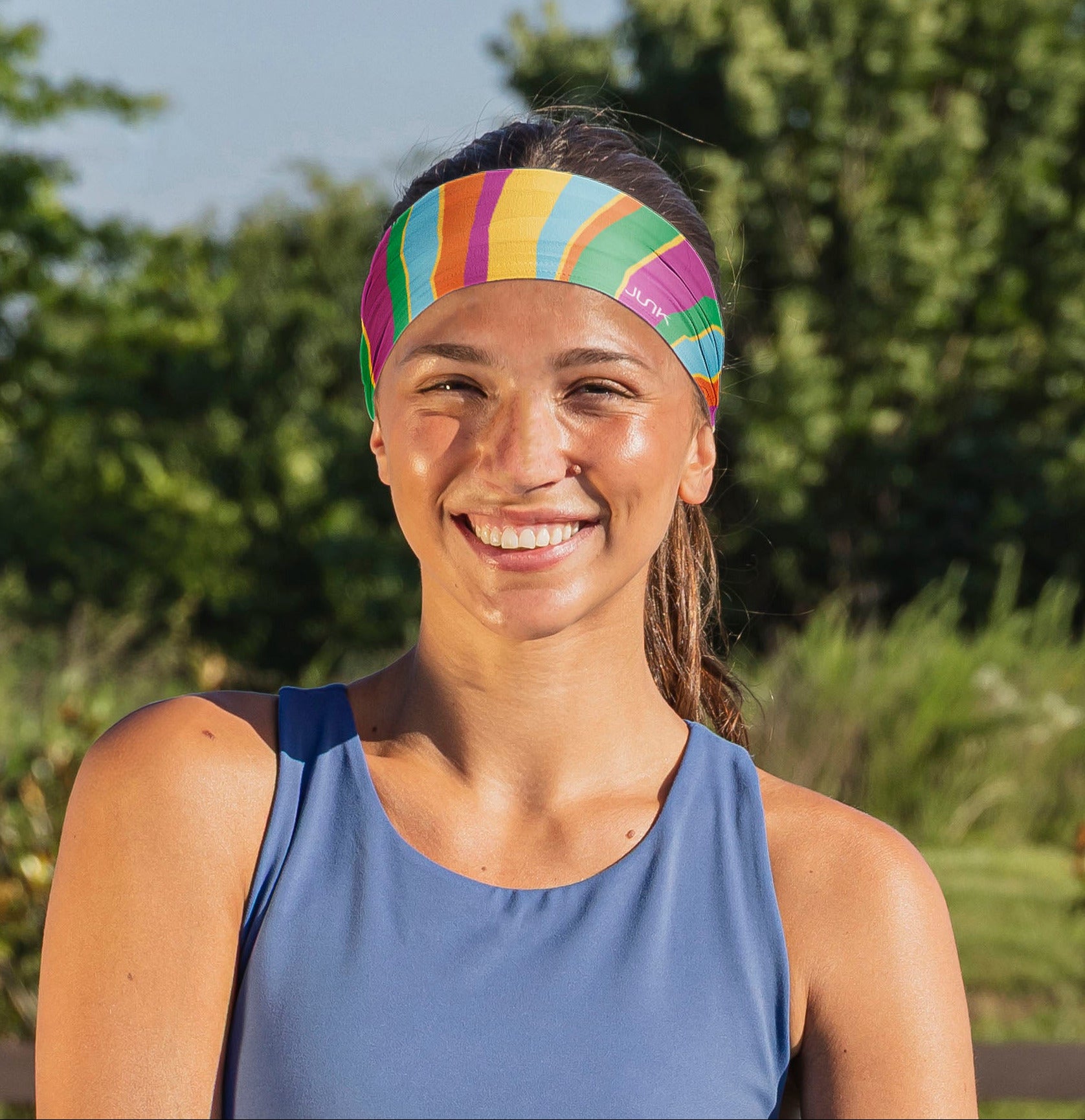 A smiling woman outdoors wearing a colorful striped athletic headband and a blue sleeveless top, with blurred green trees in the background.