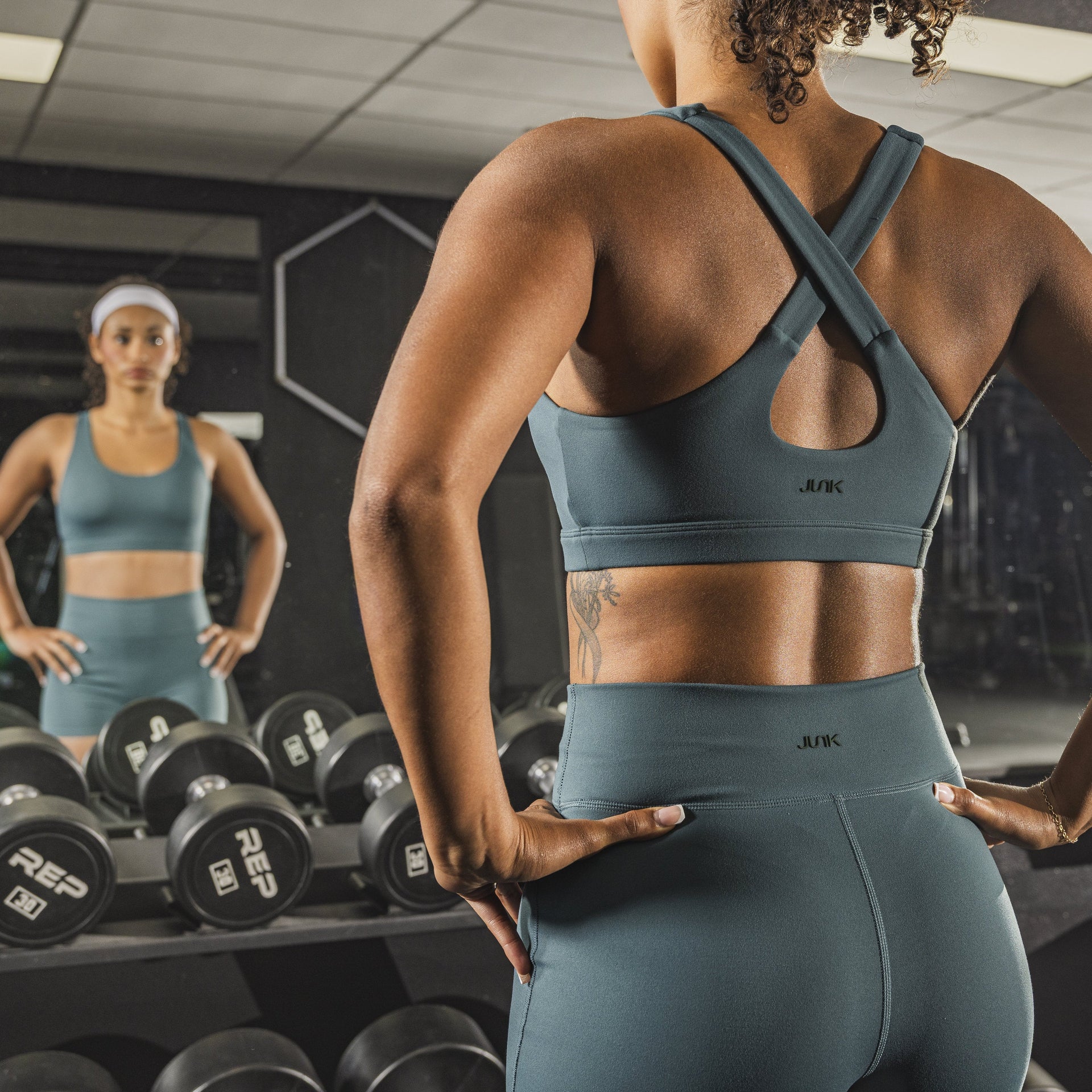 Woman in a sports bra and leggings standing in a gym, looking at herself in the mirror.