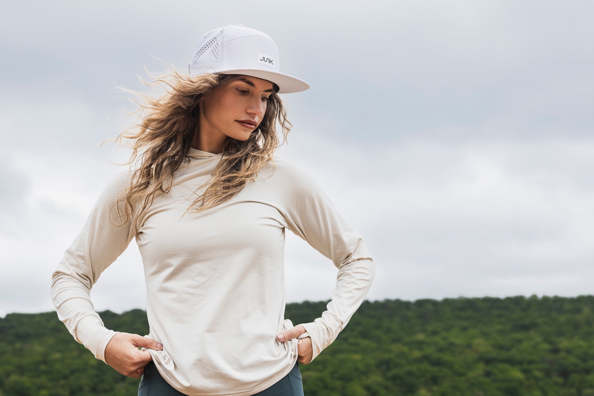 Woman wearing a white long-sleeve shirt and cap outdoors with greenery in the background