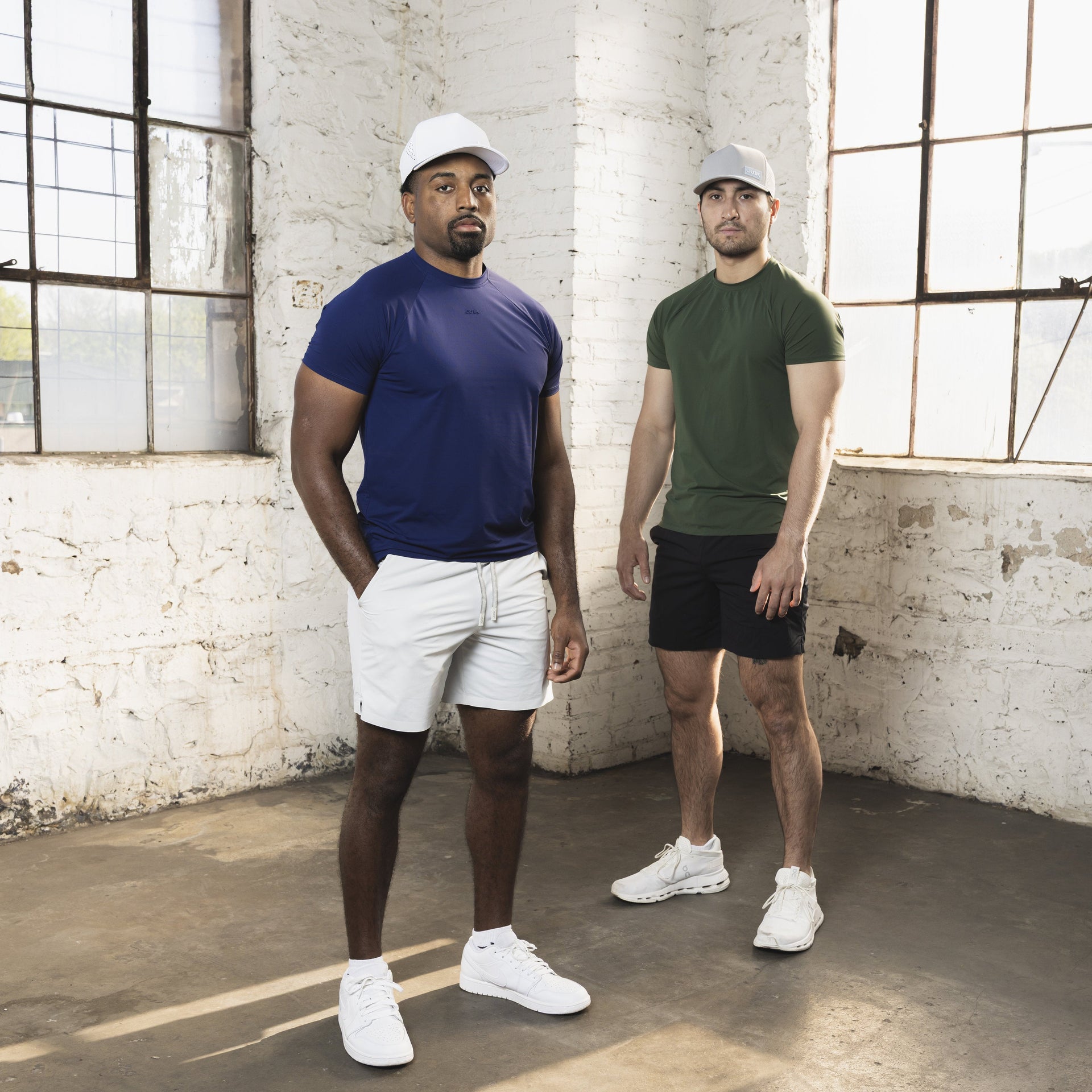 Two men standing in a room with large windows and white brick walls.