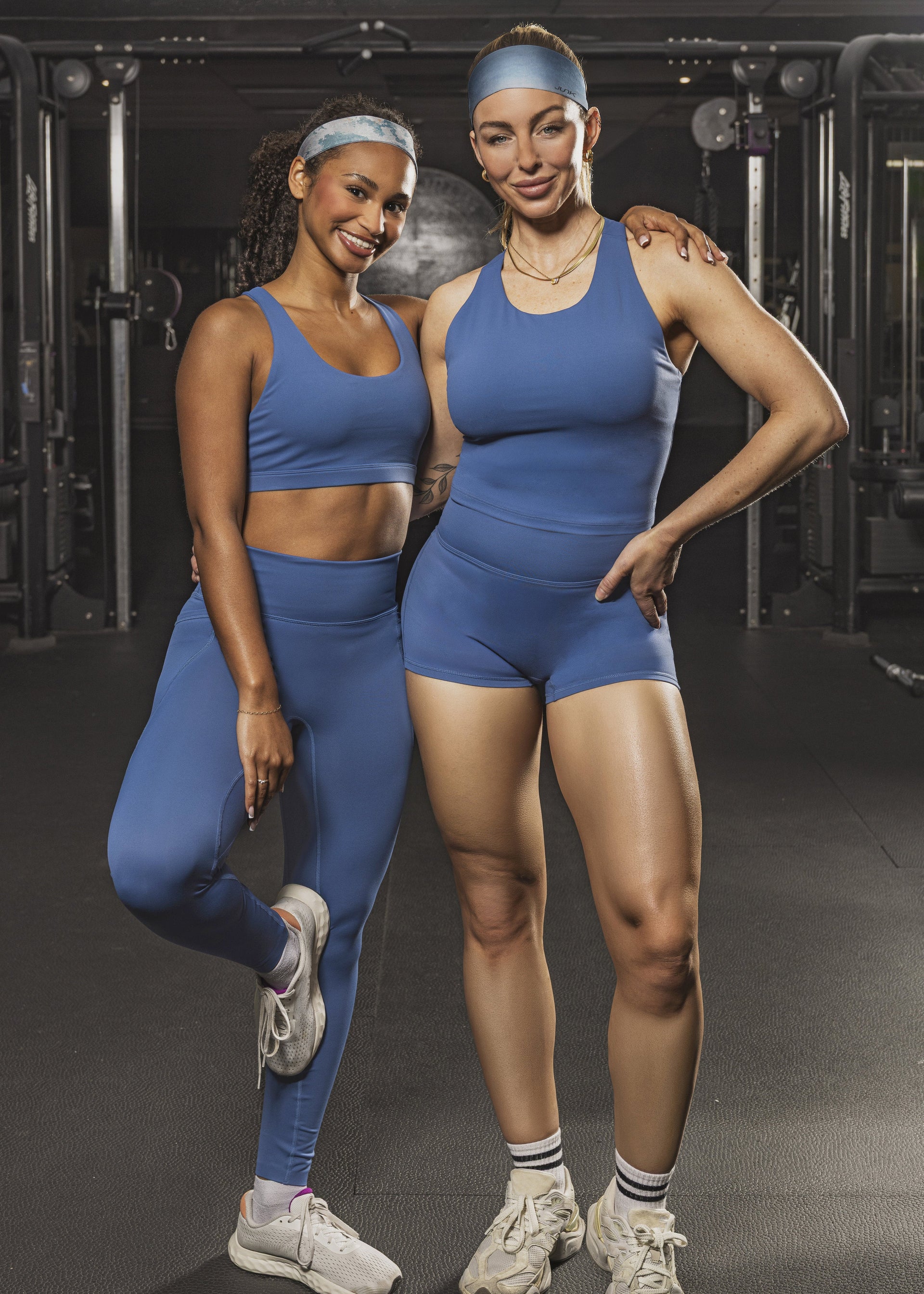 Two women posing together in a gym, both wearing matching blue athletic wear including sports bras, leggings, and shorts, with white sneakers.