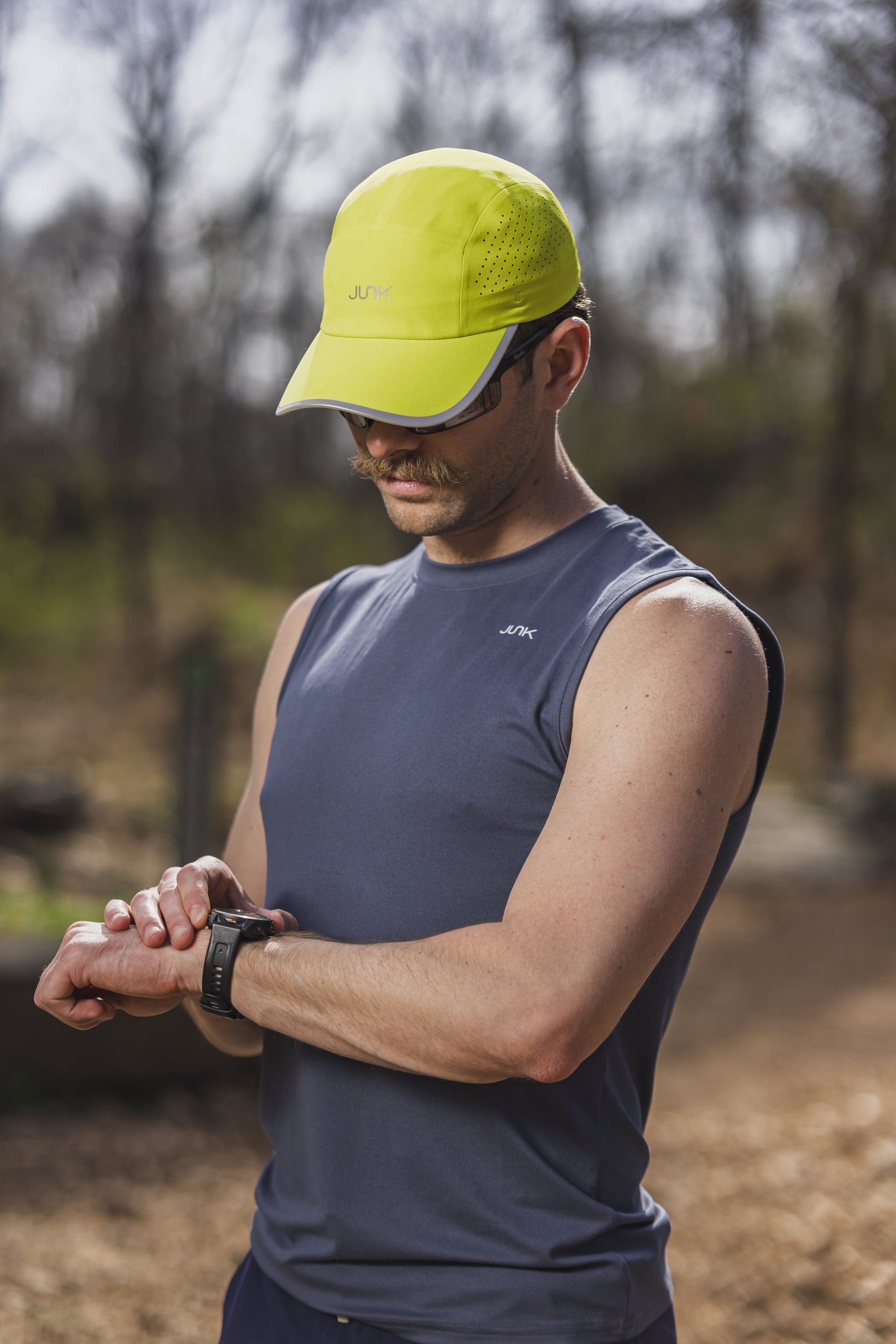 A man outdoors wearing a bright yellow athletic cap and a dark gray sleeveless top, looking down at a smartwatch on his wrist. The background is a blurred natural setting with trees.